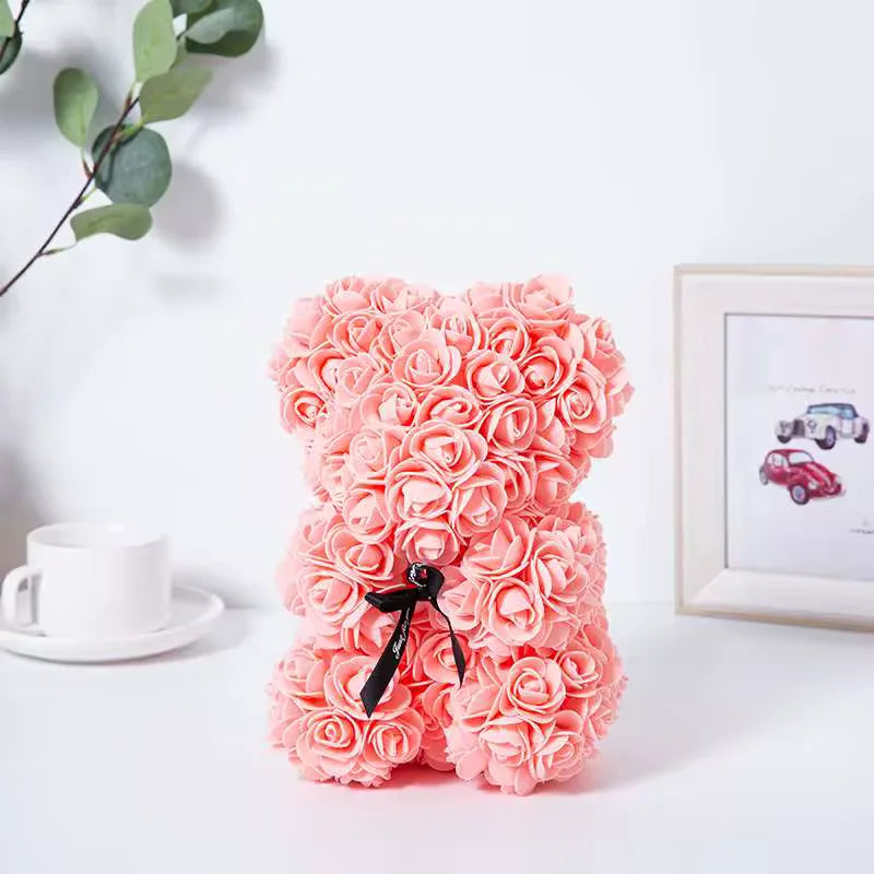 Ursinho de rosas cor rosa com laço preto ao lado de xícara branca e de um quadro